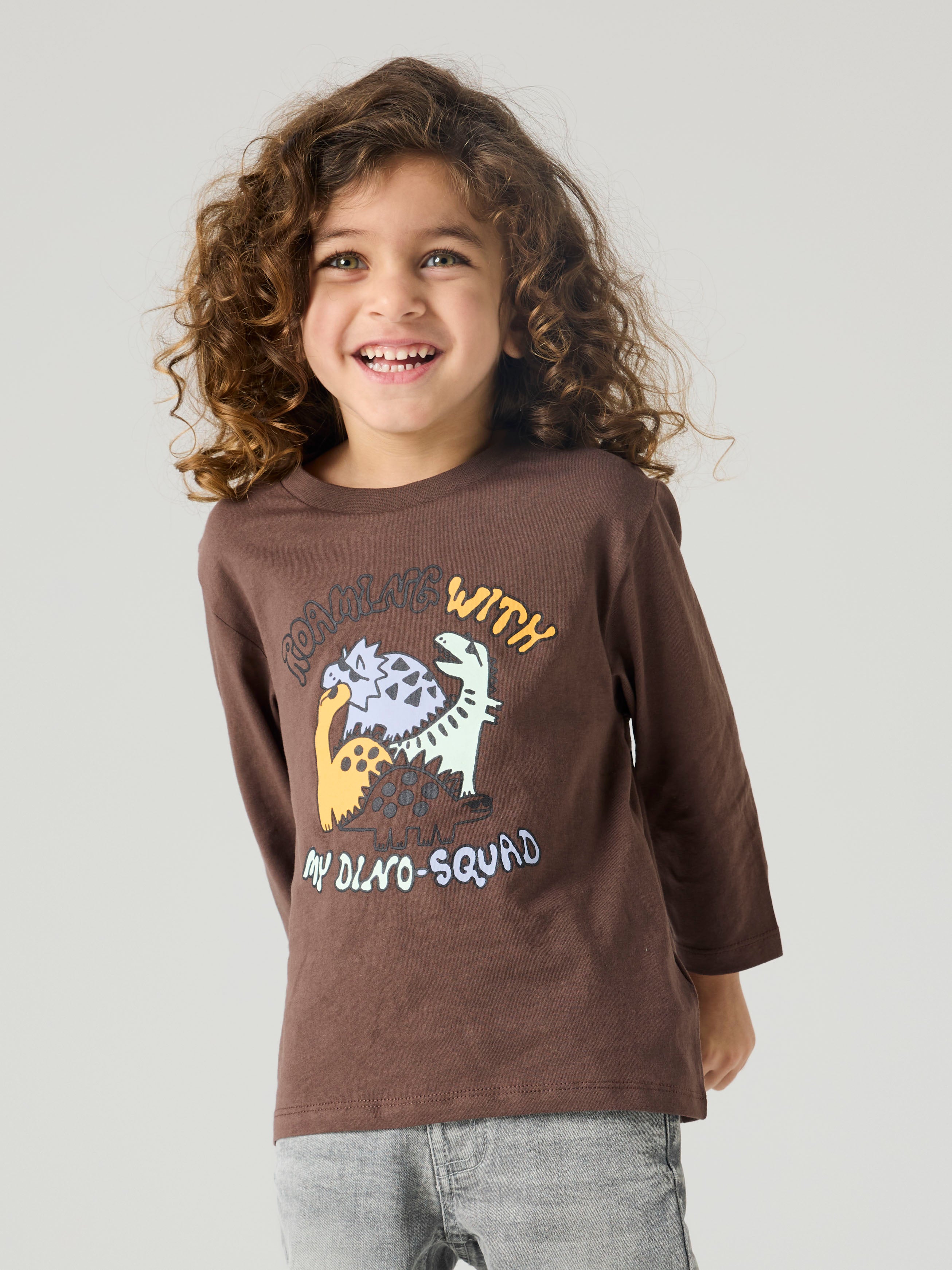 Child wearing a brown long-sleeve shirt with dinosaur graphics and text on a plain background