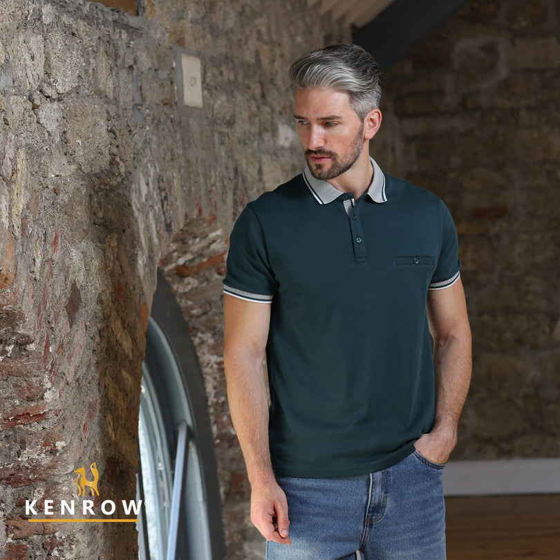 Man wearing a dark green polo shirt and jeans standing against a stone wall with 'KENROW' branding.