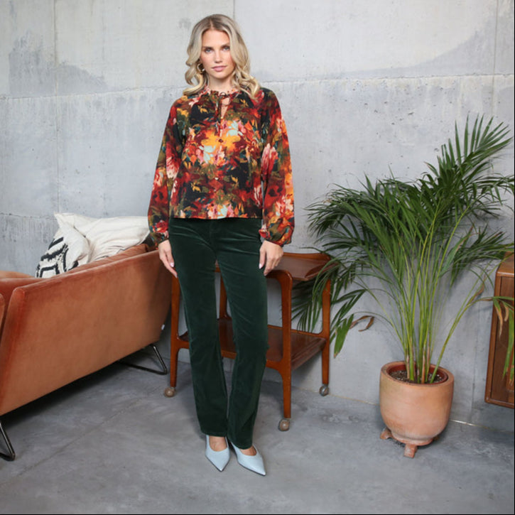 Woman in a floral blouse and green pants standing in a room with a plant and furniture.