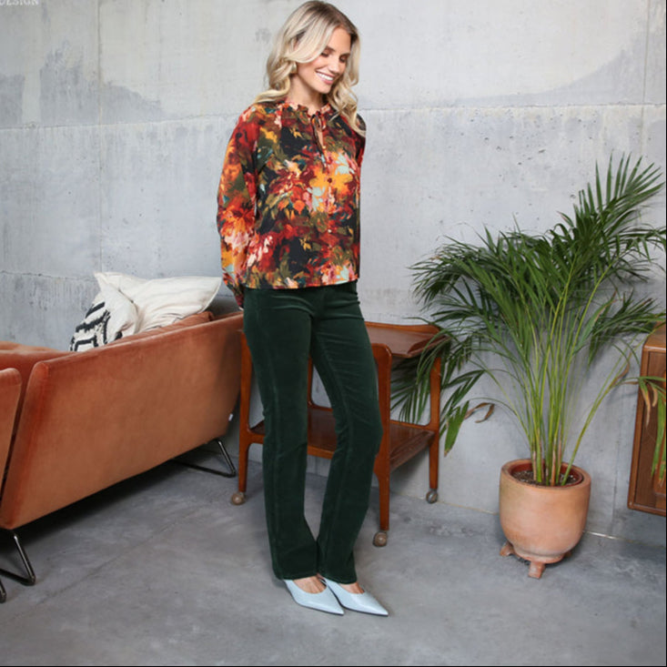 Woman in a colorful floral blouse and green pants standing in a modern living room.