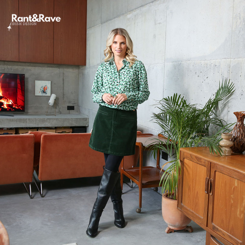 Woman in a green patterned shirt and dark green skirt standing in a modern living room with 'Rant&Rave' branding.