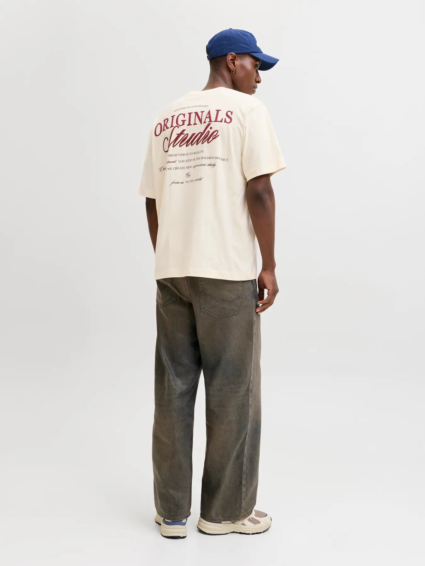 Person wearing a beige 'Originals Studio' t-shirt and blue cap on a white background