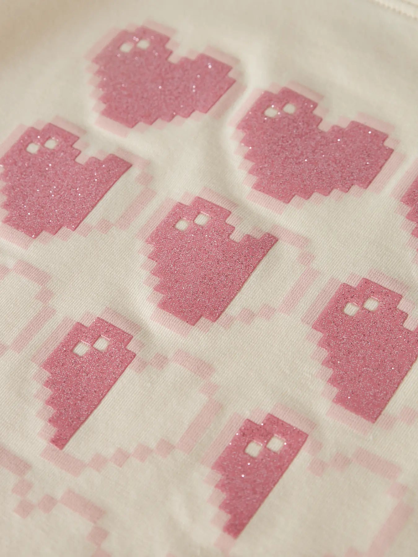 A close-up of a light cream fabric printed with pink pixelated ghost motifs, featuring a subtle glitter texture.