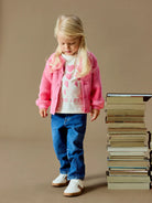 A young blonde girl wearing a pink cardigan over a white heart-print t-shirt, blue jeans, and white sneakers, standing next to a tall stack of books against a solid beige background.