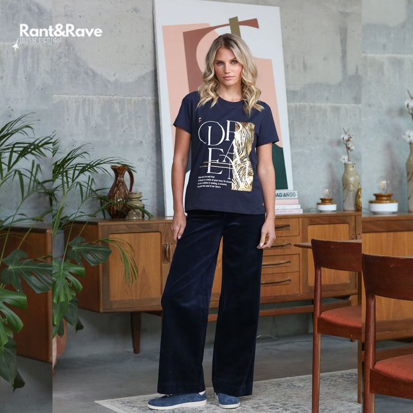 Woman wearing a navy blue t-shirt with gold design and navy wide cord pants in a room with wooden furniture and plants.