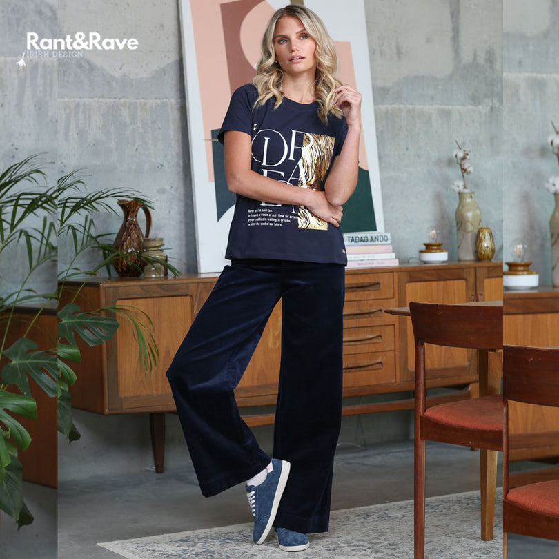 Woman wearing a navy t-shirt with gold text and navy pants in a room with wooden furniture and plants.