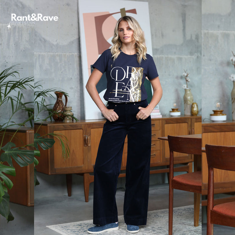 Woman wearing a graphic t-shirt and dark pants in a room with wooden furniture and plants.