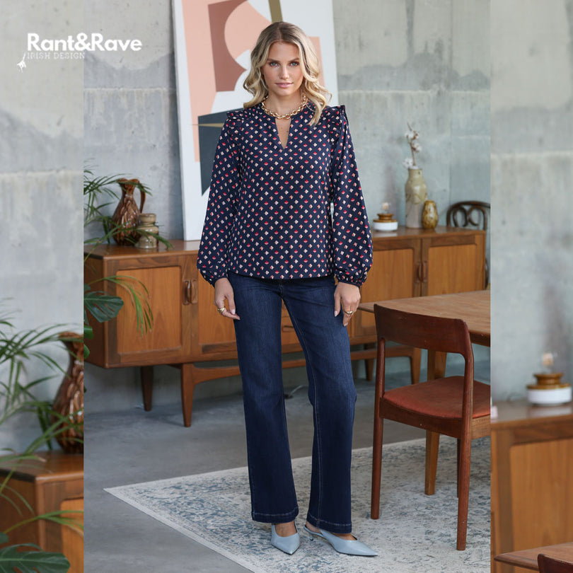 Woman wearing a patterned blouse and jeans standing in a room with wooden furniture and decor.