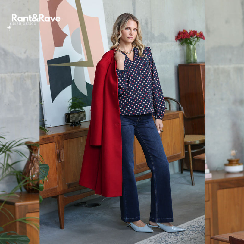Woman in a polka dot blouse and red coat standing in a room with wooden furniture and abstract art.
