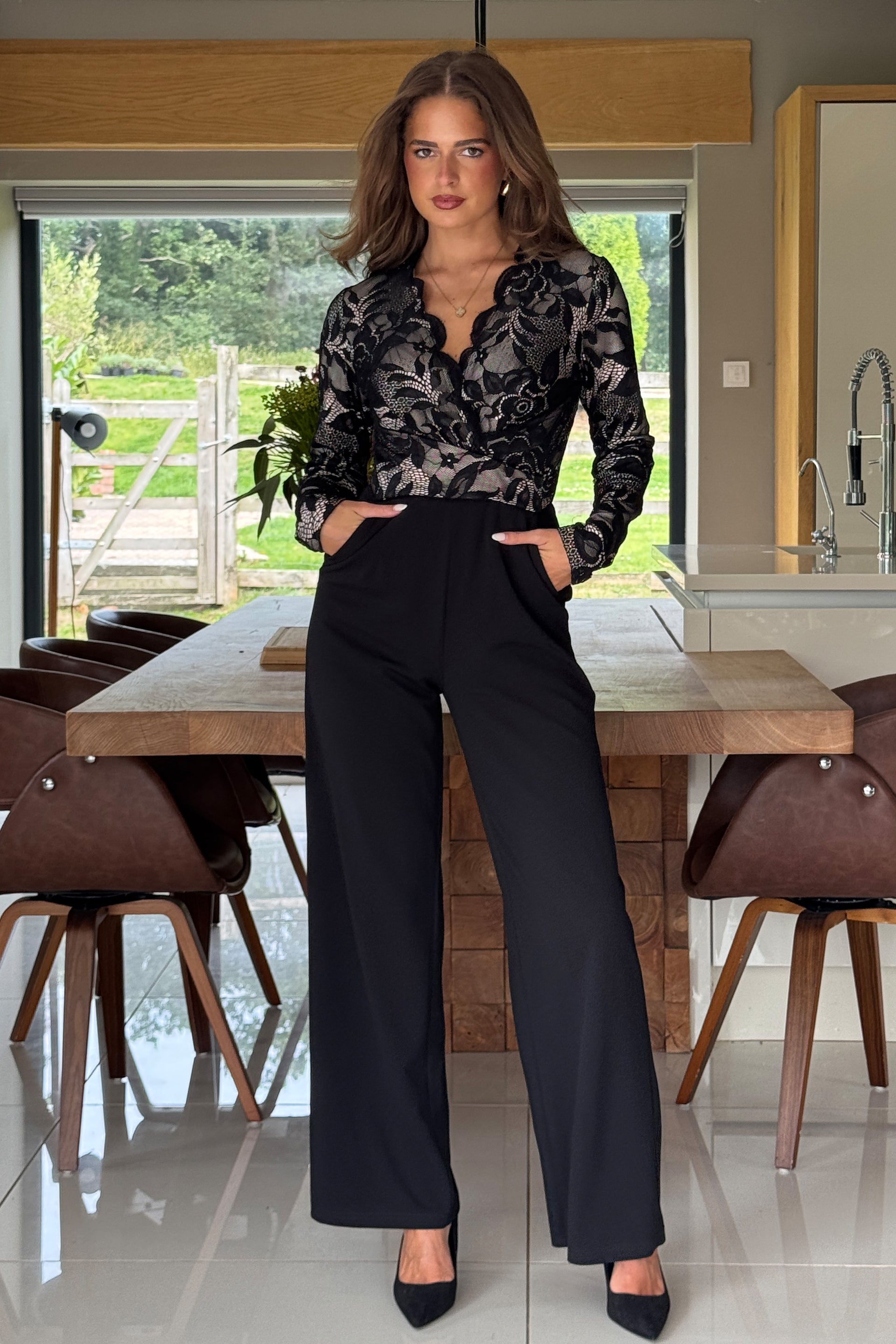 Woman in a lace top jumpsuit with black bottoms standing at a dining table in a modern kitchen.