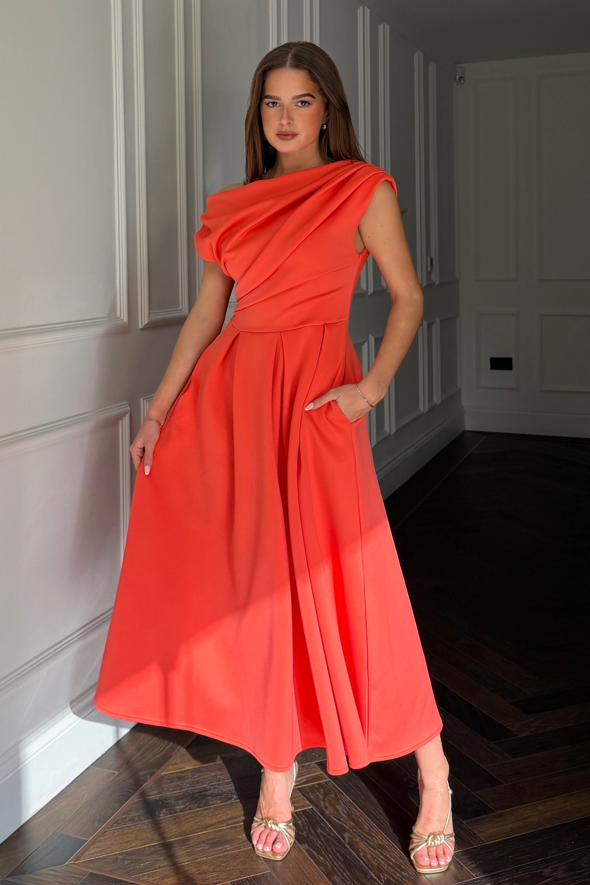 Woman in a coral red dress standing in a room with wooden flooring and white walls.