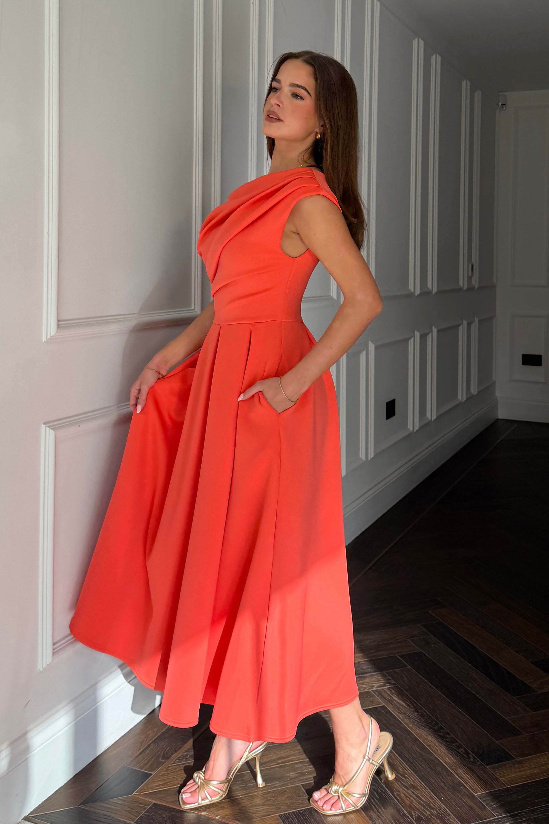 Woman in a coral red dress standing in a hallway with white paneled walls.