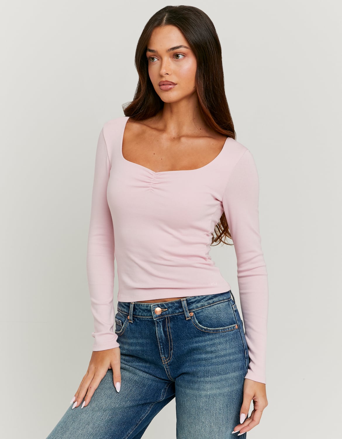 Woman wearing a pink long-sleeve top and blue jeans on a plain background