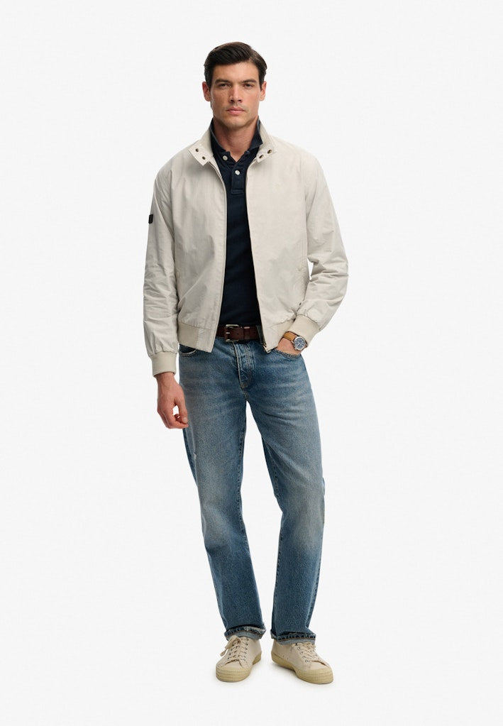 Man wearing a grey jacket, dark shirt, blue jeans, and beige shoes on a white background