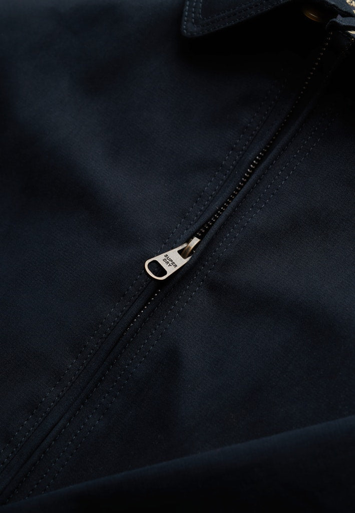 Close-up of a zipper on a dark navy harrington jacket with a brand logo.