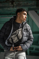 Person wearing a black and gray jacket with a bag, standing against a green wall.