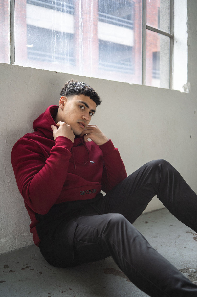 Person wearing a red hoodie sitting against a wall with large windows.