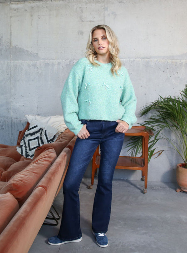 Woman in a light green sweater and dark jeans standing in a modern living room with a brown sofa and concrete wall.
