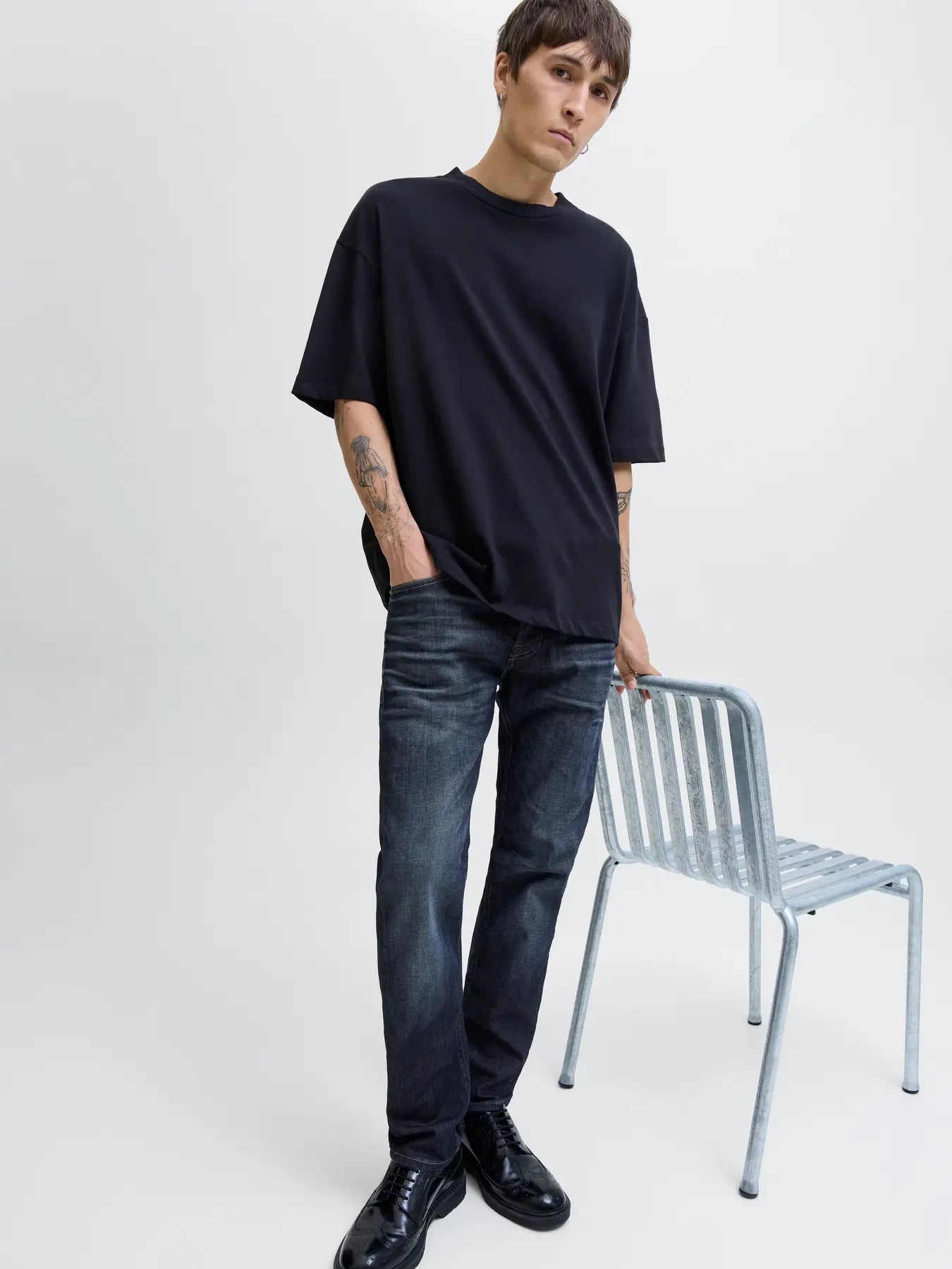 Person wearing a black oversized t-shirt and dark jeans standing next to a chair on a white background