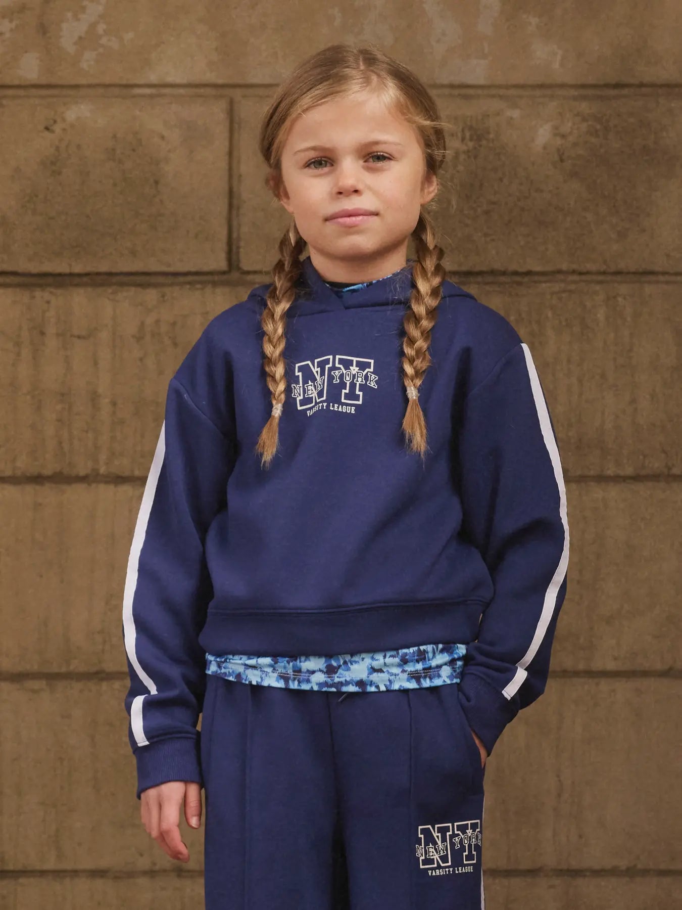 Young girl wearing a navy blue hoodie with a logo against a brown brick wall.