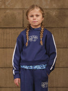 Young girl wearing a navy blue hoodie with a logo against a brown brick wall.