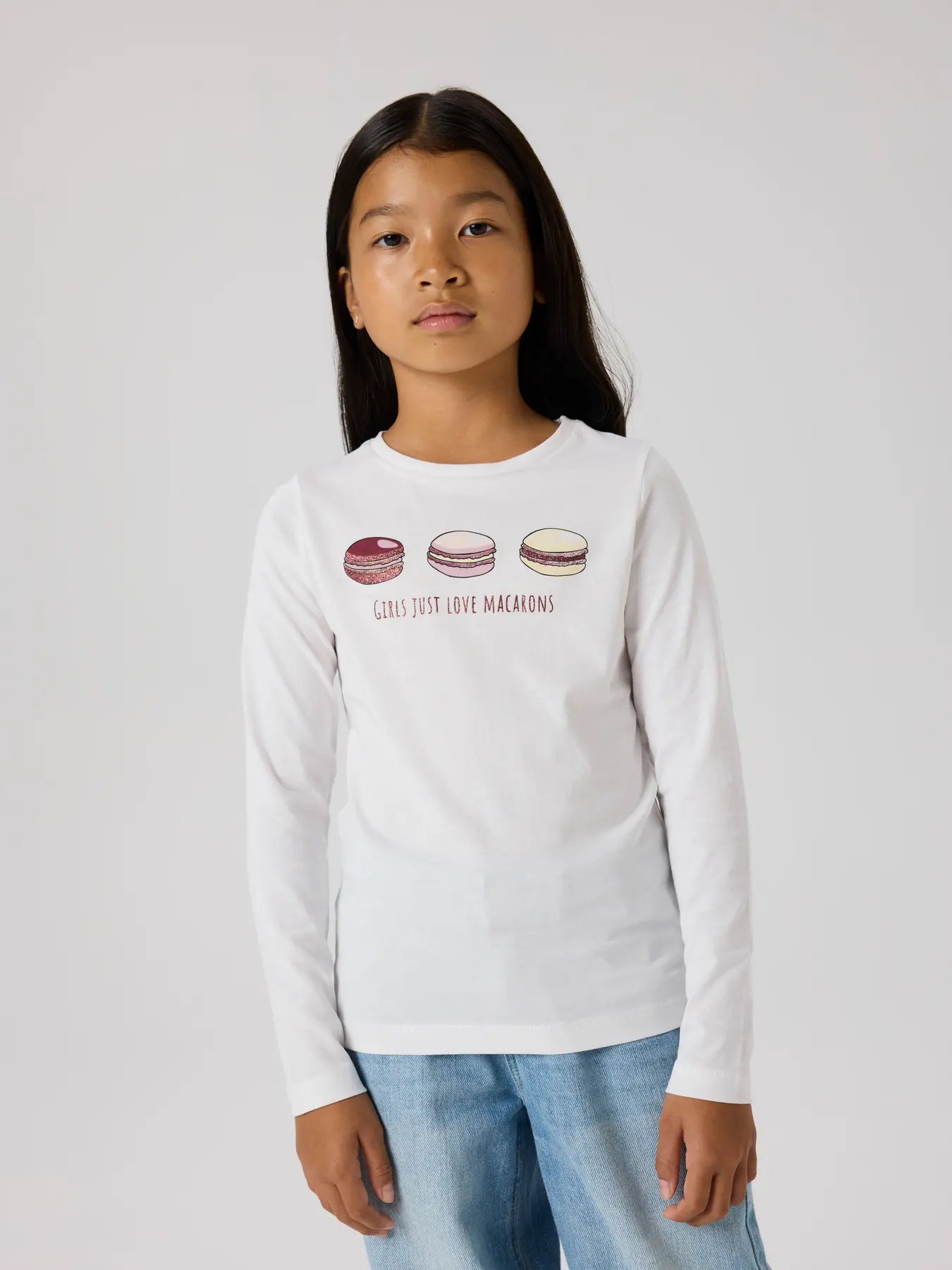 Child wearing a white long-sleeve shirt with macaron graphics and text on a plain background