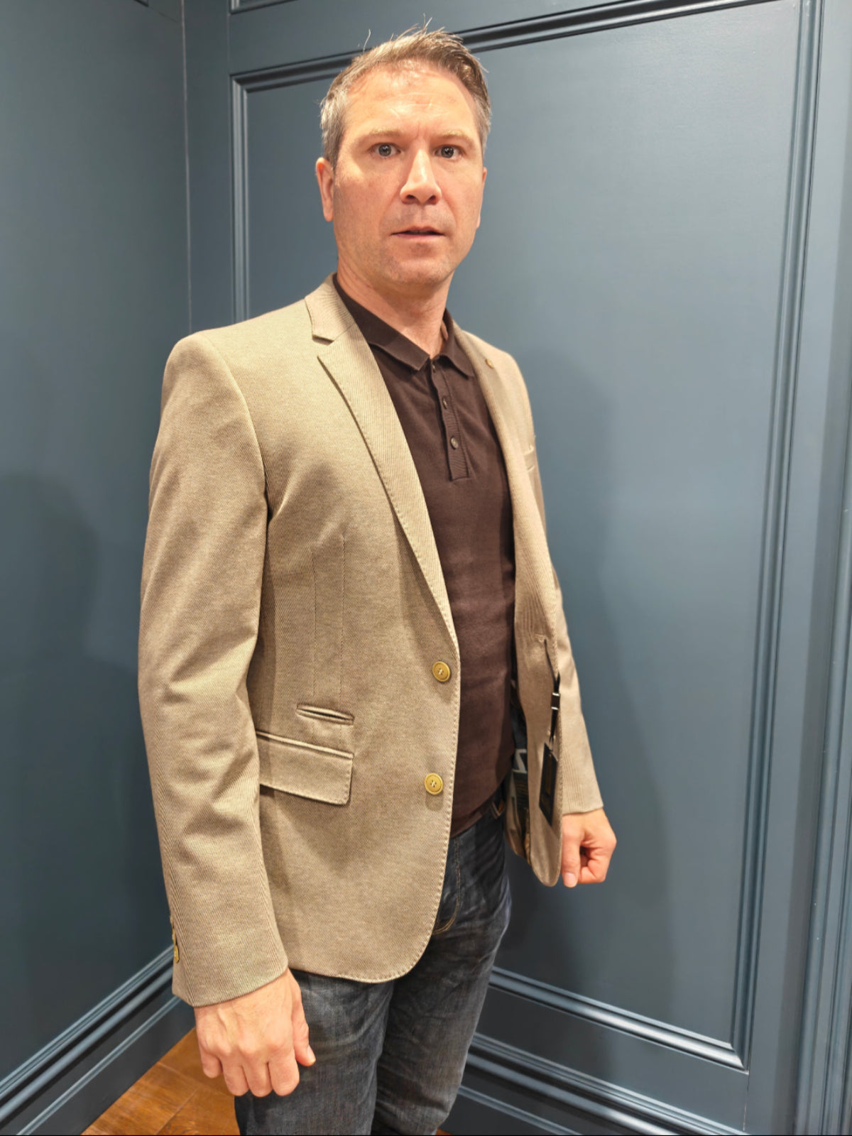 Man wearing a beige blazer over a brown shirt against a blue paneled wall.