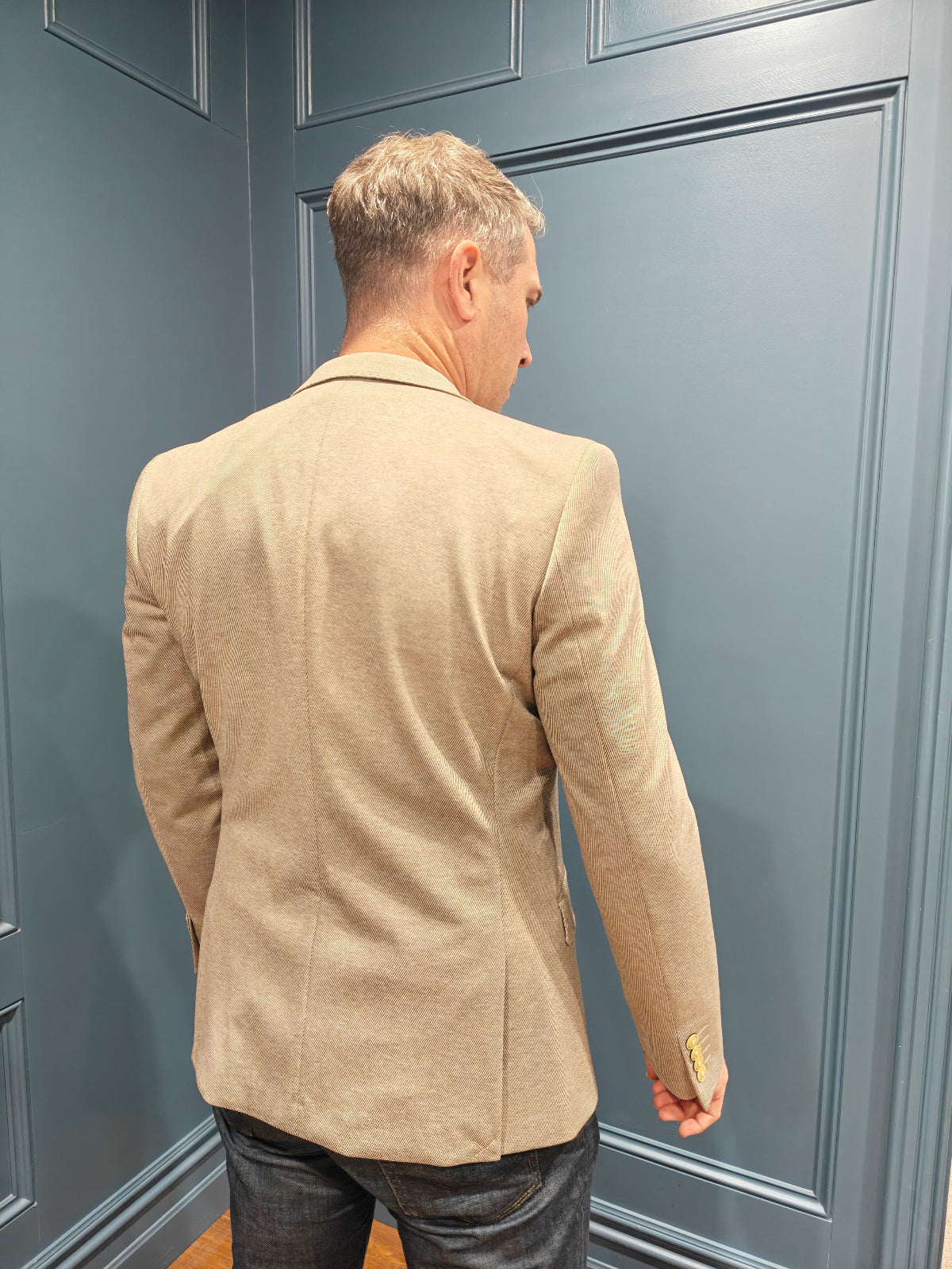 Man wearing a beige blazer standing against a gray paneled wall.