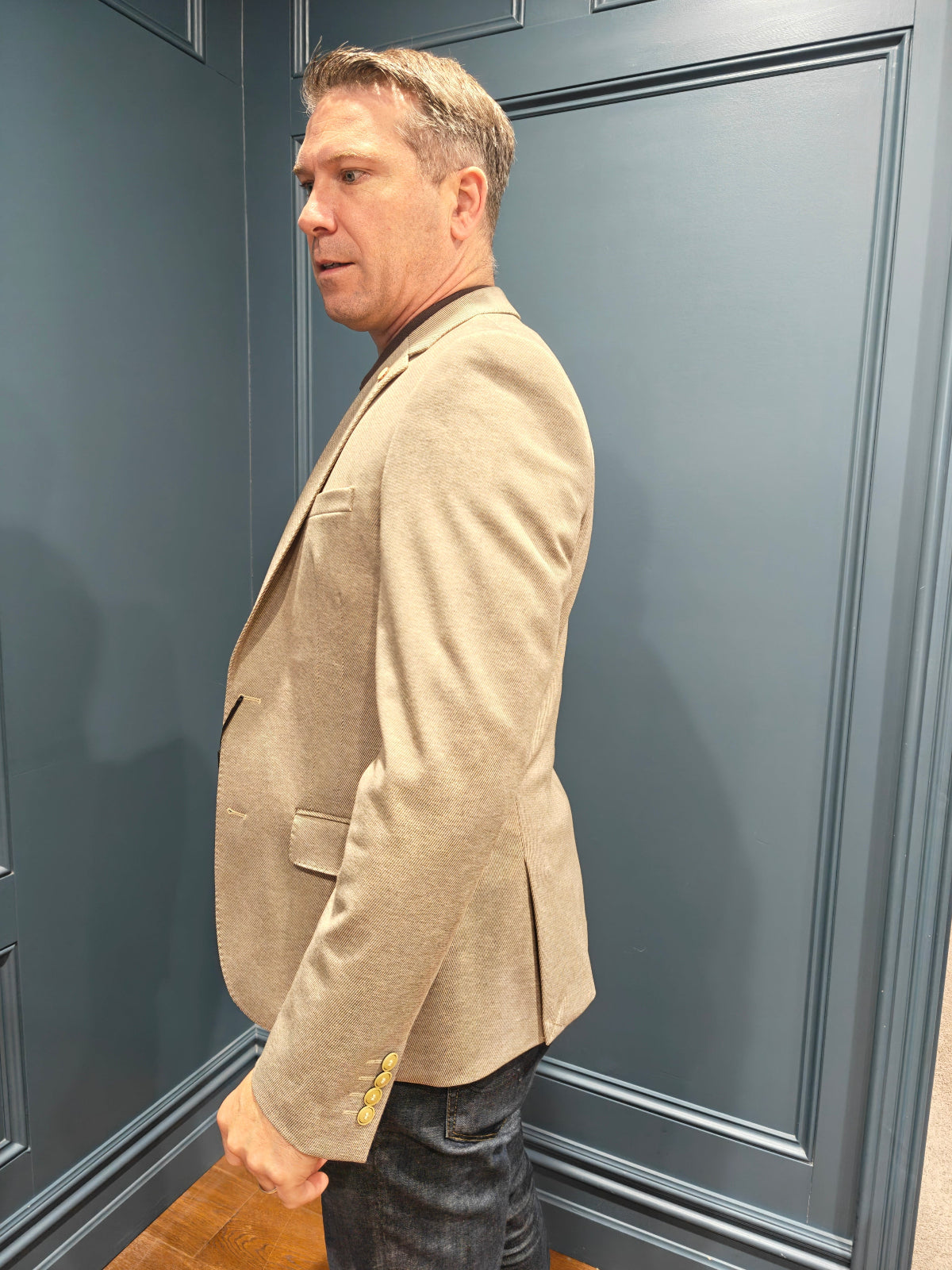 Man wearing a beige blazer standing against a gray wall.