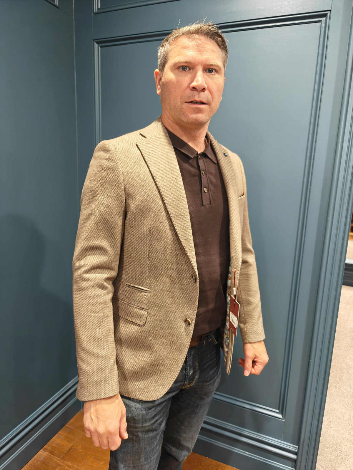 Man wearing a beige blazer and brown shirt standing against a blue wall.