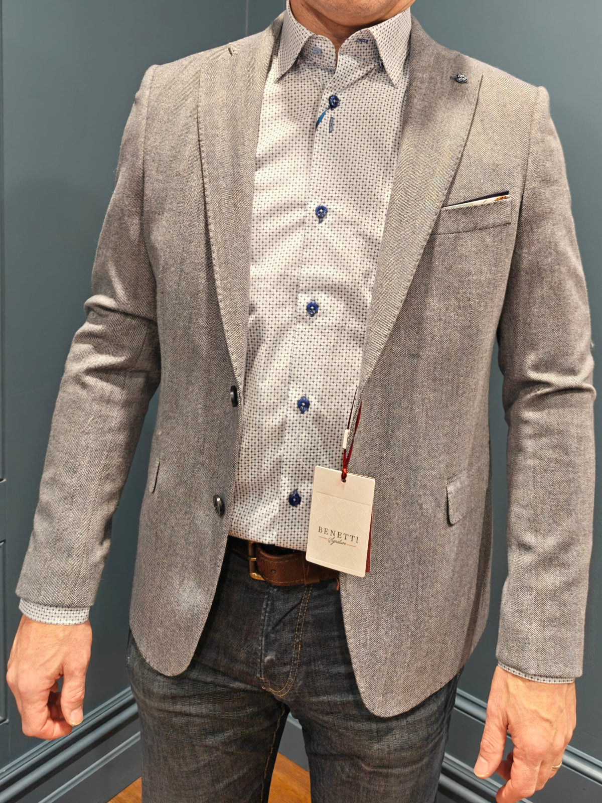 Man wearing a gray blazer over a patterned shirt with a visible tag against a gray wall.