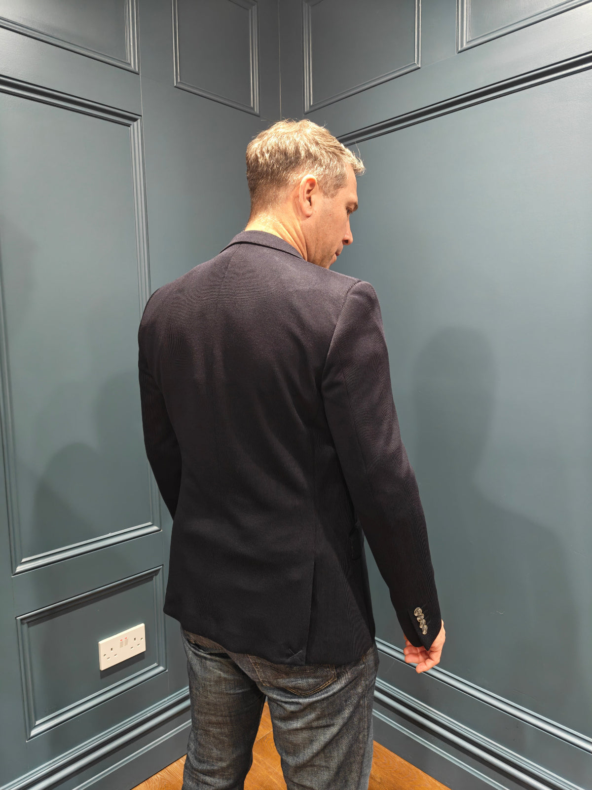 Man wearing a dark blazer and gray pants standing against a gray paneled wall.
