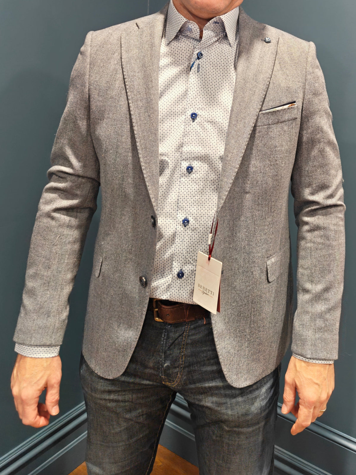 Man wearing a gray blazer and dark jeans standing against a dark blue wall.