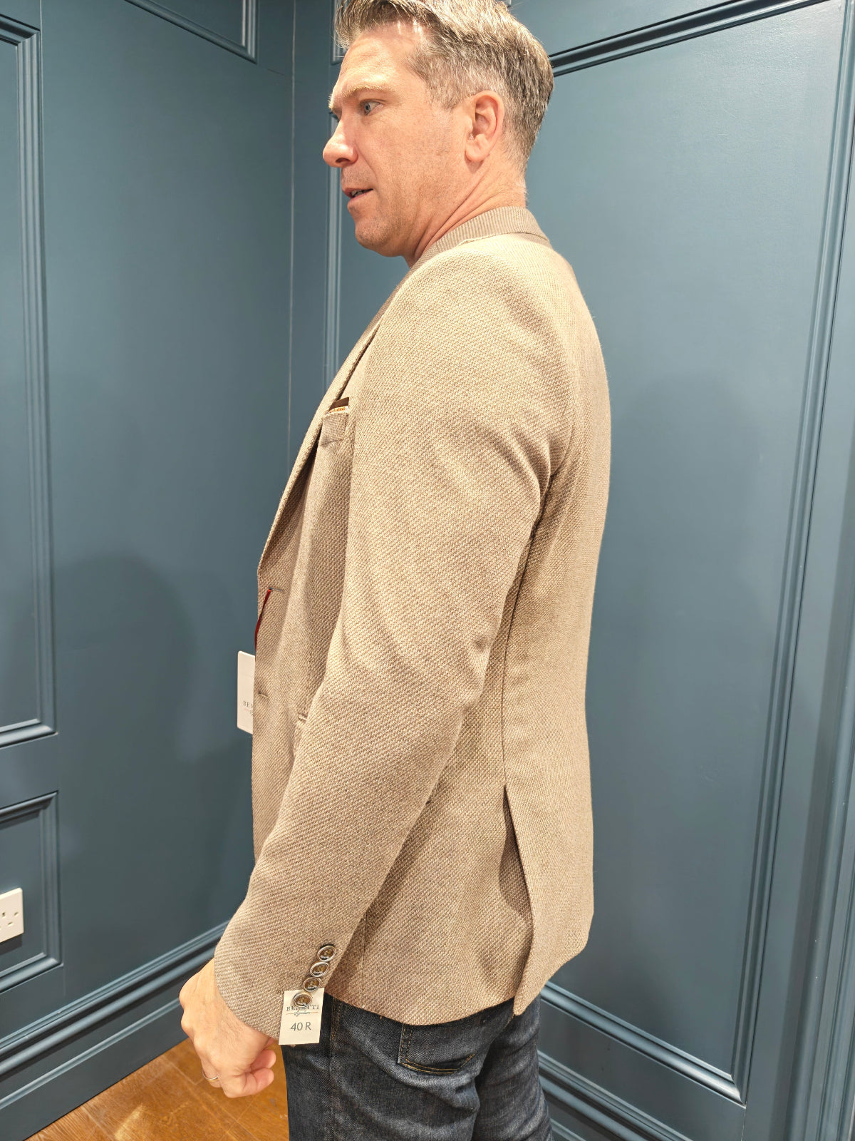 Man wearing a beige blazer with a visible tag against a blue wall.