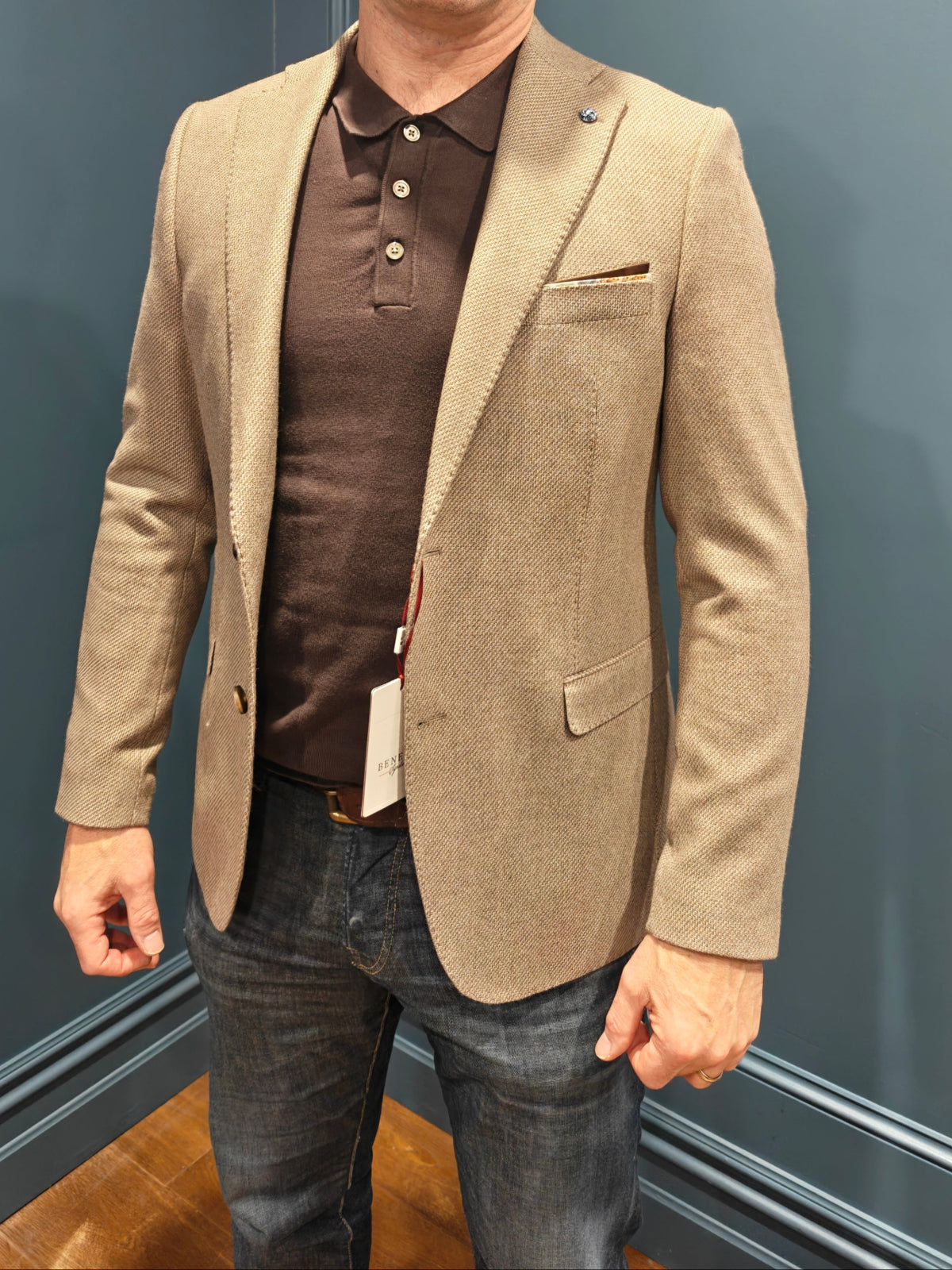 Man wearing a beige blazer over a brown shirt against a blue wall.