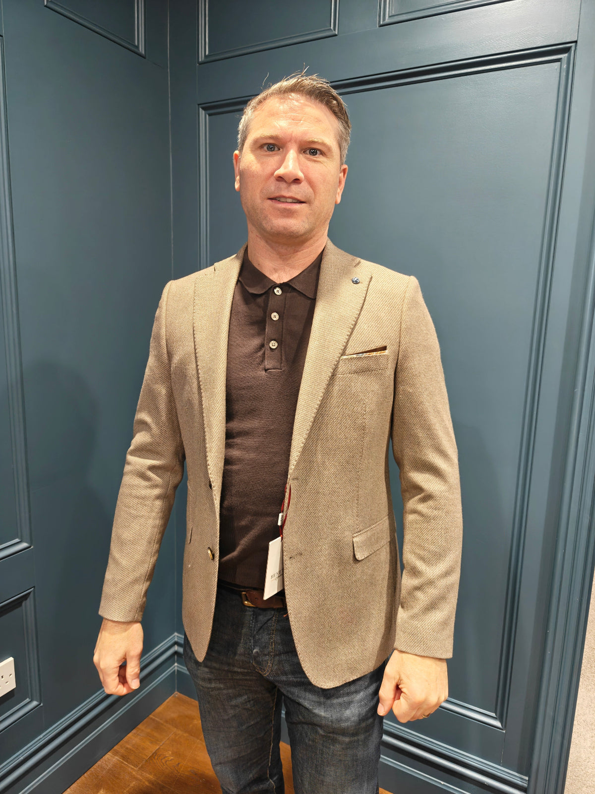 Man wearing a beige blazer over a brown shirt against a blue paneled wall.