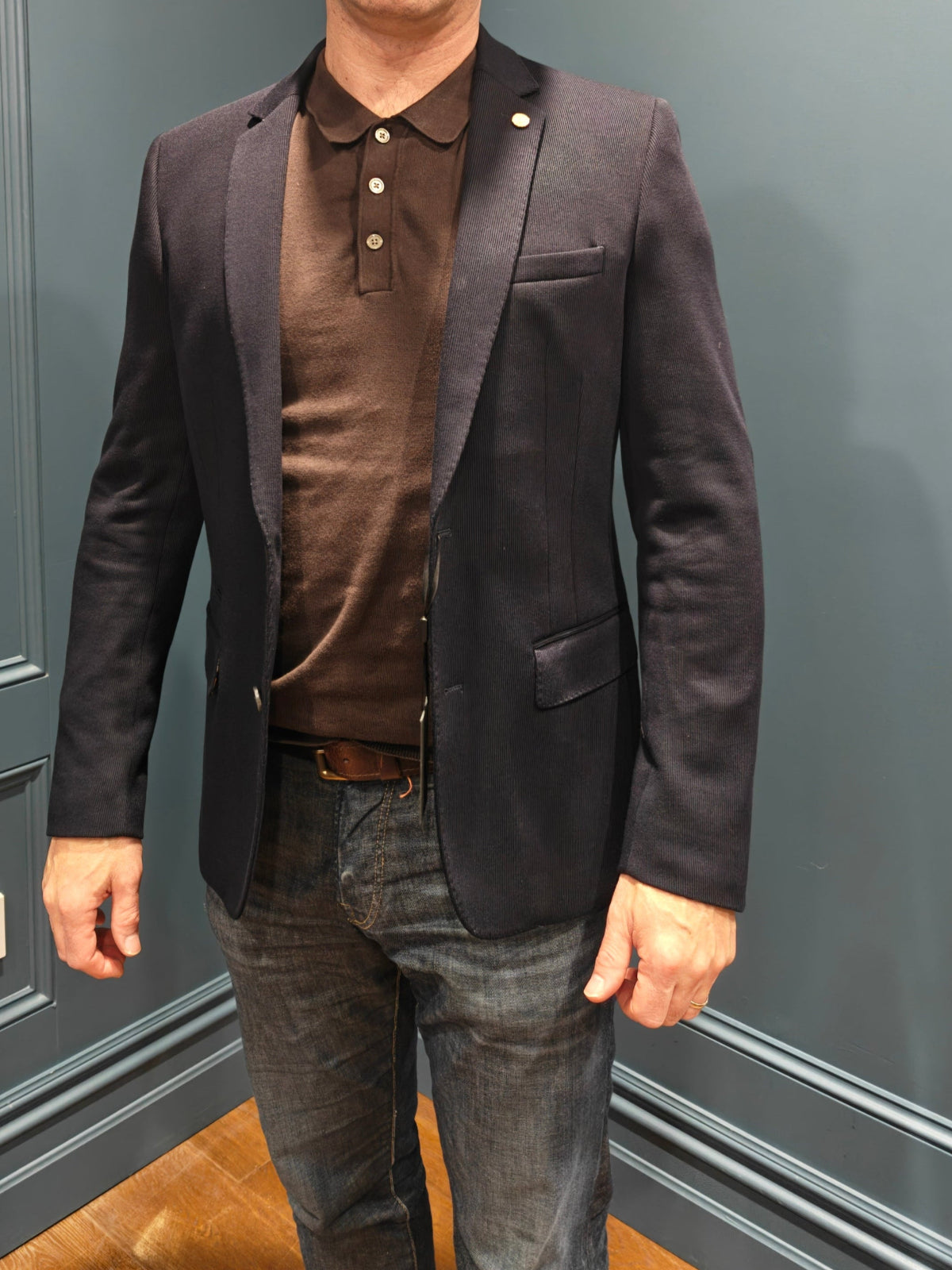 Man wearing a dark blazer over a brown shirt and jeans standing against a gray paneled wall.