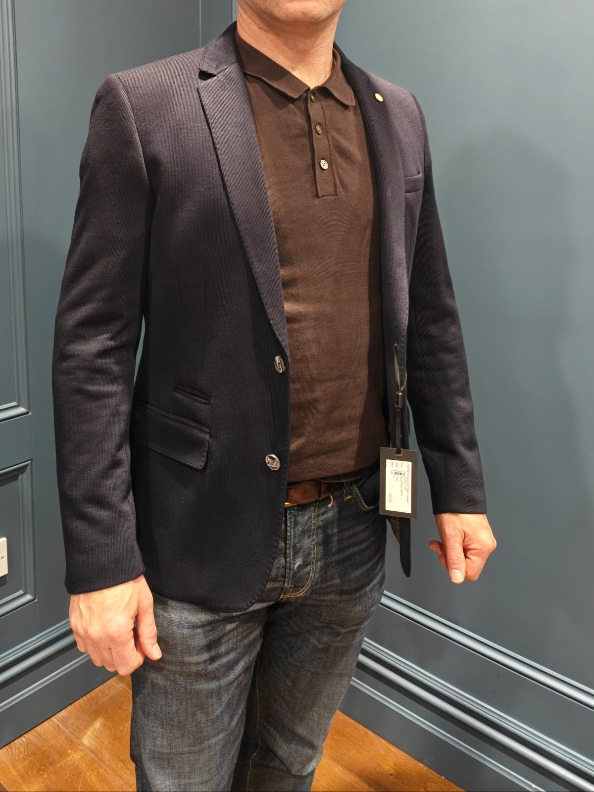 Man wearing a dark blazer over a brown shirt and jeans standing against a gray paneled wall.