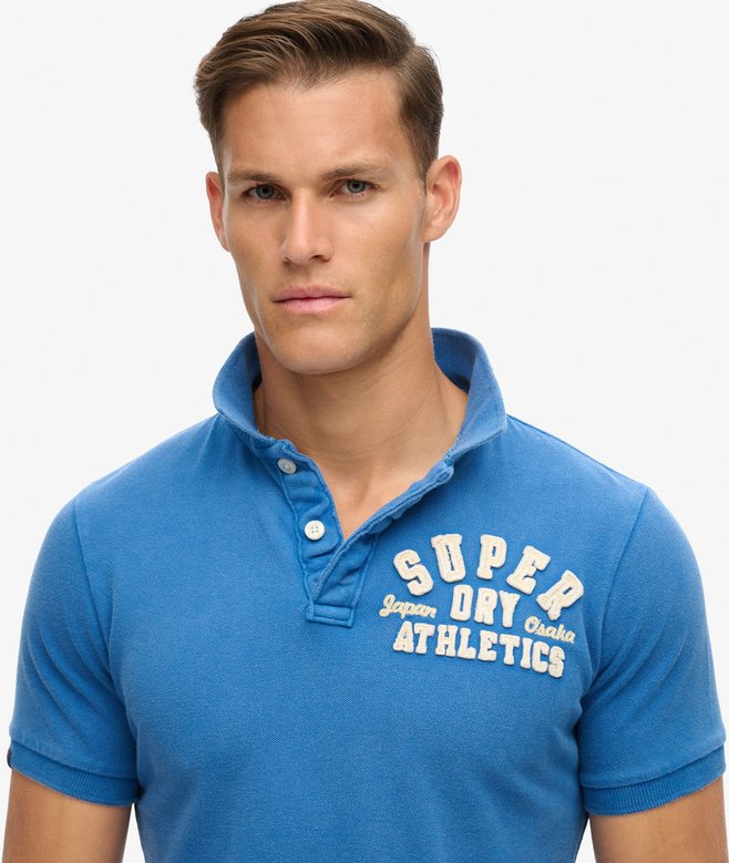 Man wearing a blue 'Super Dry Athletics' polo shirt on a white background