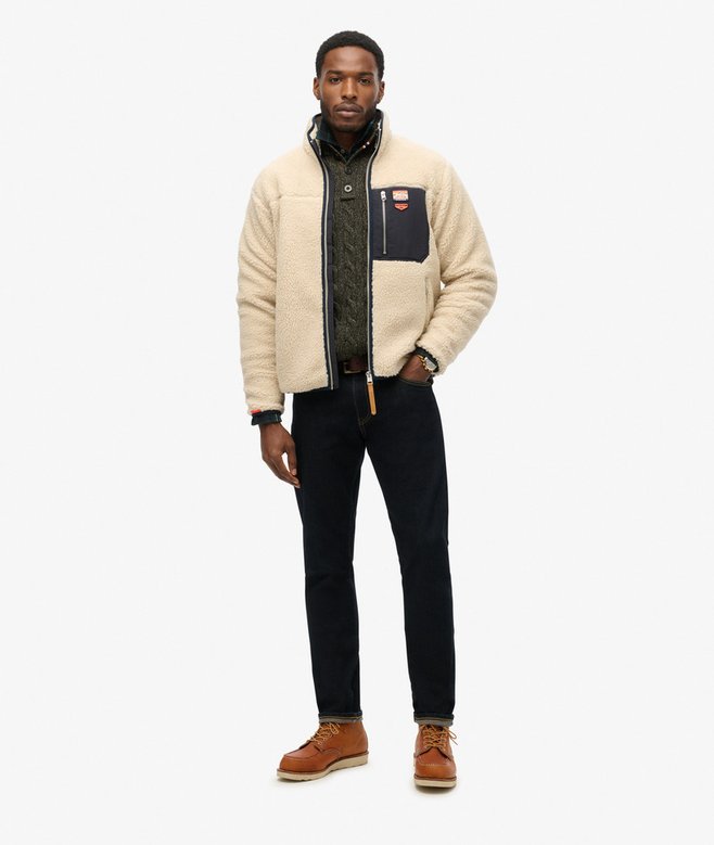 Man wearing a beige jacket with a black patch, dark pants, and brown shoes on a white background