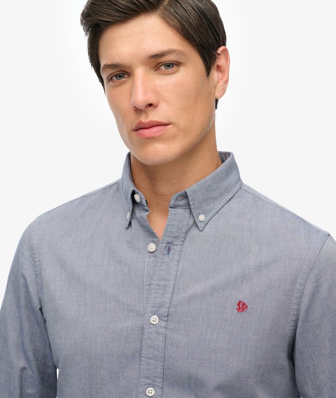 Man wearing a bluebutton-up shirt with a small red logo on a white background