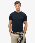 Man wearing a navy blue t-shirt and beige pants with a plaid shirt over his shoulder on a white background
