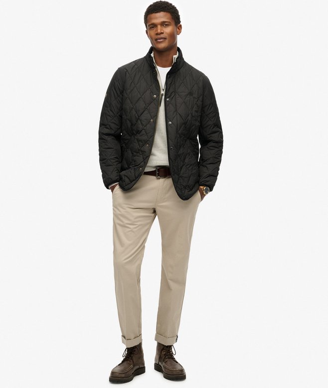 Man wearing a black quilted jacket, white shirt, beige pants, and brown shoes on a white background