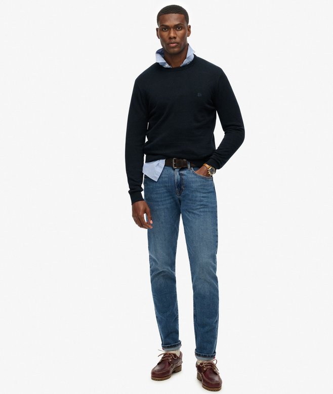 Man wearing a navy sweater, blue jeans, and brown shoes on a white background