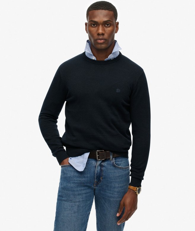 Man wearing a navy sweater over a collared shirt with blue jeans on a white background