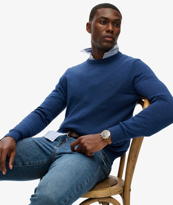 Man wearing a blue sweater and jeans sitting on a wooden chair against a white background