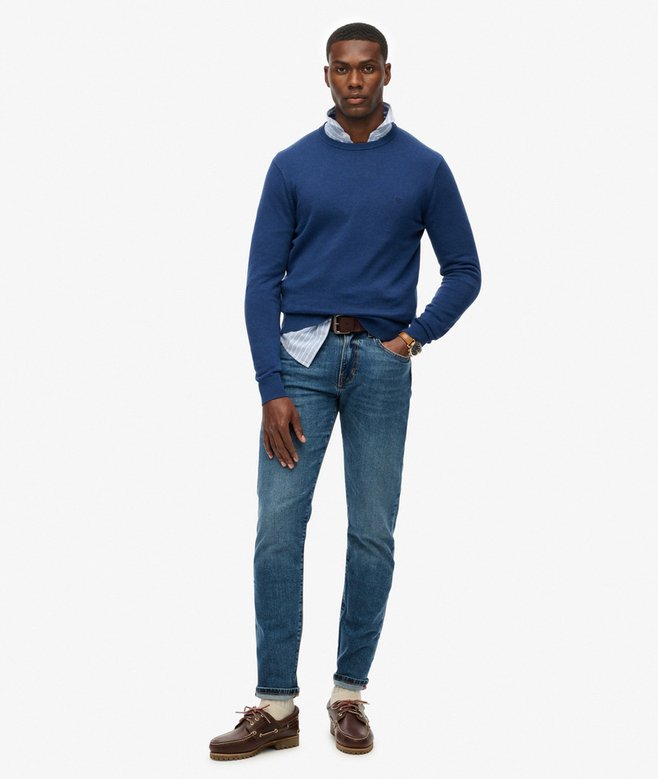 Man wearing a blue sweater, white shirt, and jeans on a white background