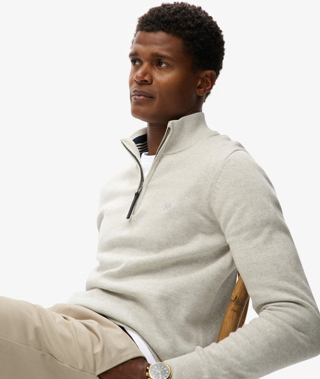 Man wearing a light gray half-zip sweater sitting on a chair with a white background