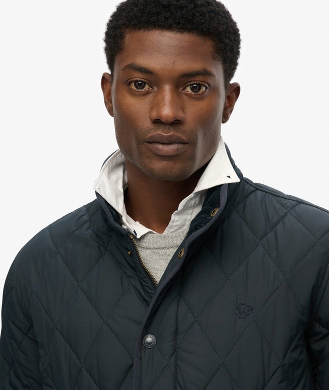 Man wearing a navy quilted jacket over a white shirt with a gray sweater underneath, on a plain background.
