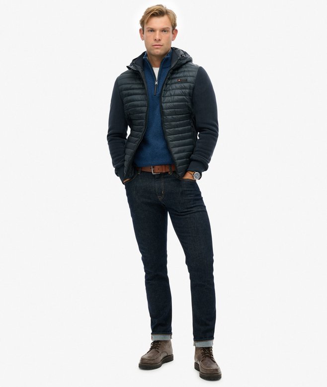 Man wearing a dark blue puffer jacket, navy sweater, jeans, and brown shoes on a white background